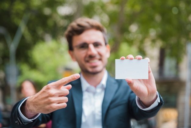 Porträt eines defokussierten jungen Geschäftsmannes, der seinen Finger in Richtung zur weißen Visitenkarte zeigt