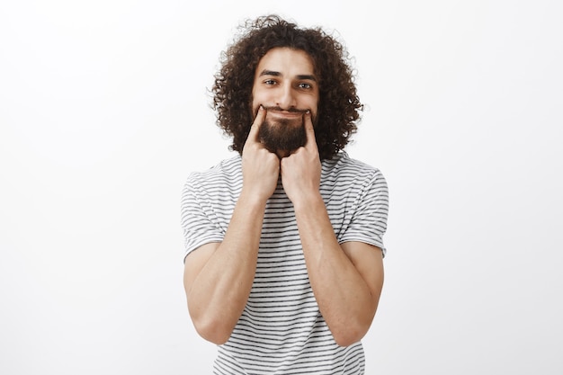 Porträt eines depressiven unglücklichen gutaussehenden hispanischen bärtigen Kerls mit lockigem Haar, das mit Zeigefingern ein Lächeln zieht und versucht, positiv auszusehen, während er sich verärgert und traurig fühlt