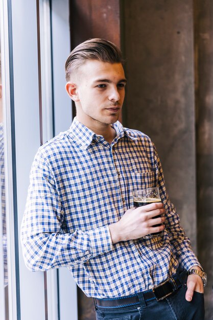 Porträt eines durchdachten jungen Mannes, der am Fenster in der Hand hält das Bierglas sich lehnt