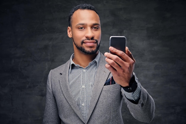 Porträt eines eleganten bärtigen schwarzen amerikanischen Mannes in einem grauen Anzug hält ein Smartphone.
