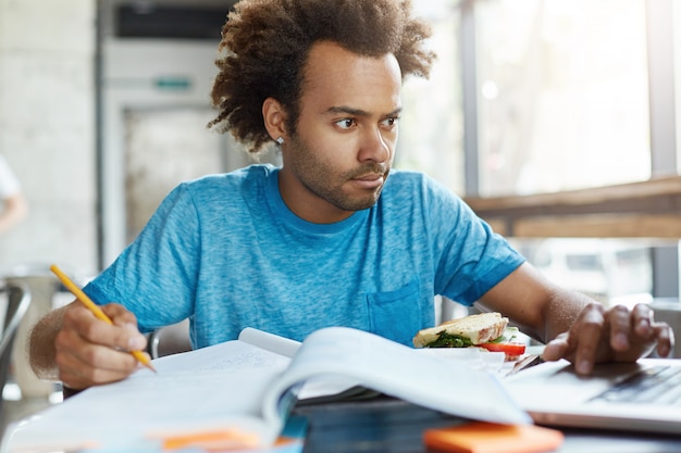 Porträt eines ernsthaften dunkelhäutigen studenten, der aufmerksam in notizbuch schaut, das notizen im schulheft schreibt, das für klassen an der universität vorbereitet, die fastfood essen. konzentrierter mann, der sehr beschäftigt ist