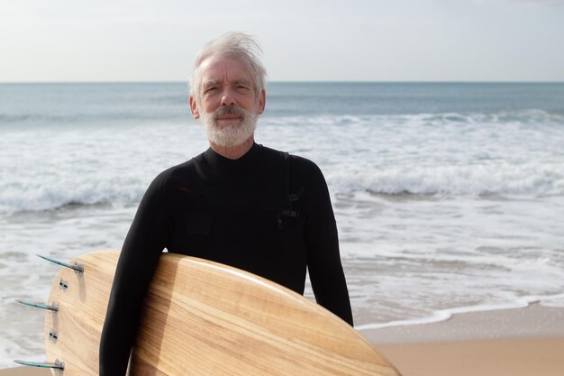 Porträt eines ernsthaften gealterten Mannes mit Surfbrett. Nachdenklicher bärtiger Mann, der am Meeresstrand steht und ein großes Holzbrett hält und in die Kamera schaut. Konzept für Gesundheit, Sport und Freizeit für ältere Menschen