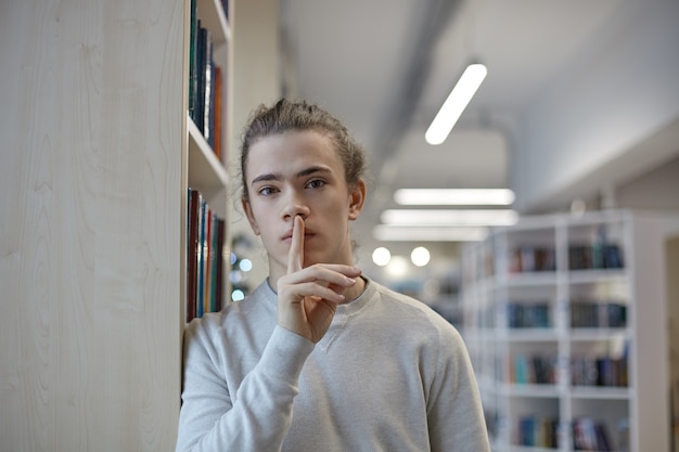 Porträt eines ernsthaften Schülers in der Freizeitkleidung, die in der Bibliothek steht und Schweigegeste zeigt