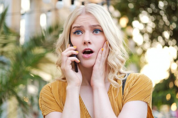 Porträt eines erstaunten blonden Mädchens, das beim Telefonieren im Freien verblüfft wegschaut