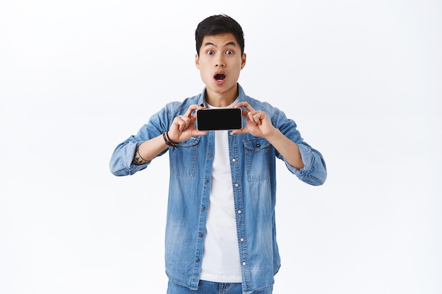 Porträt eines erstaunten jungen beeindruckten asiatischen Mannes, der einen neuen Trailer des Films auf dem Smartphone-Bildschirm zeigt, das Mobiltelefon horizontal hält, den Mund amüsiert öffnet