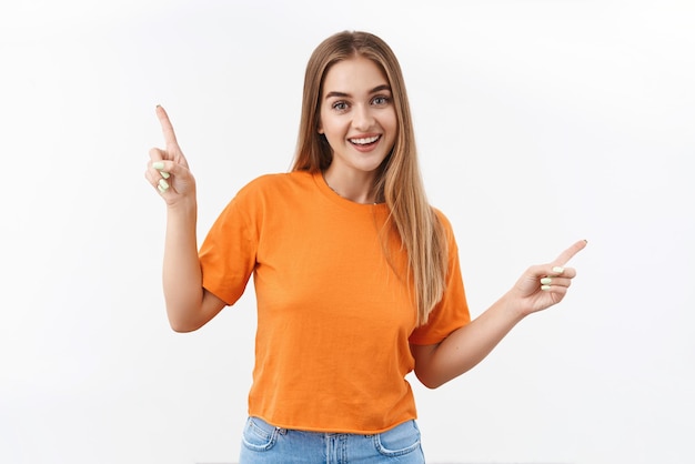 Porträt eines fröhlichen, attraktiven blonden Mädchens in orangefarbenem T-Shirt hilft bei der Auswahl und zeigt mit den Fingern seitlich, die den Weg nach links und rechts zeigen