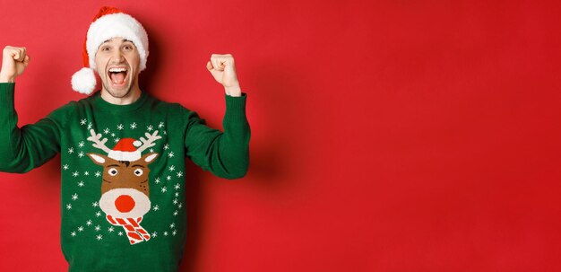 Porträt eines fröhlichen, attraktiven Mannes, der das neue Jahr feiert, einen grünen Pullover und eine Weihnachtsmütze trägt, vor Freude schreit, gewinnt oder triumphiert und auf rotem Hintergrund steht