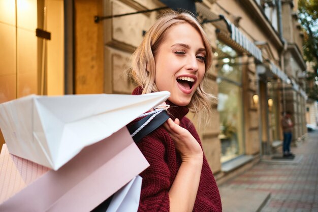 Porträt eines fröhlichen blonden Mädchens mit Einkaufstüten, das glücklich auf der Straße der Stadt blinzelt