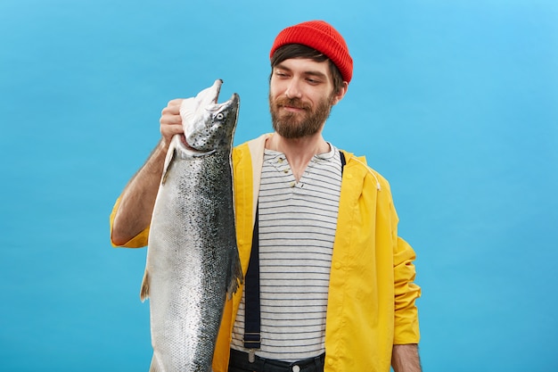 Porträt eines fröhlichen Fischers, der einen roten Hut, eine gelbe Jacke und einen Overall trägt und mit erfreutem Ausdruck auf seinen Fang schaut, der stolz ist.