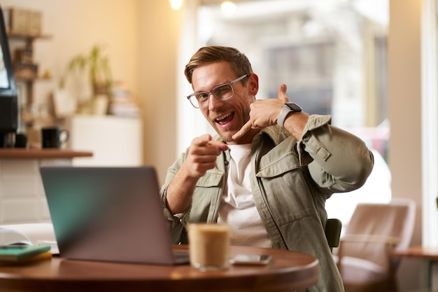 Porträt eines fröhlichen, flirtende Mannes mit Brille, der mit einem Laptop in einem Café sitzt und einen Telefonanruf zeigt