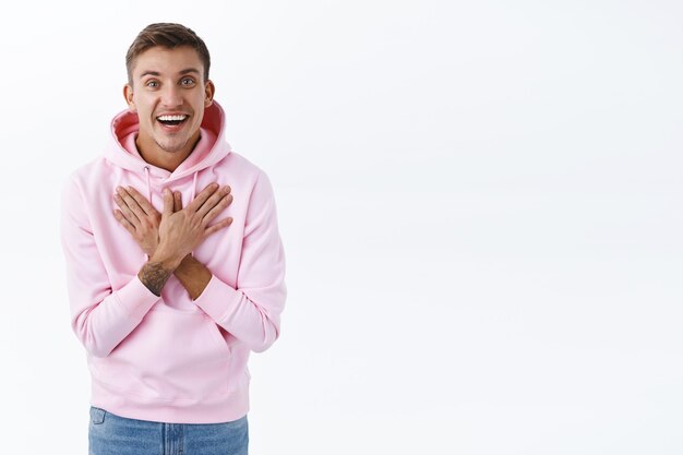 Porträt eines fröhlichen, glücklichen blonden Kerls in rosa Hoodie, die Hände auf der Brust halten dankbar oder erfreut, großartige Neuigkeiten zu hören, und lächelt und freut sich über gute Leistungen