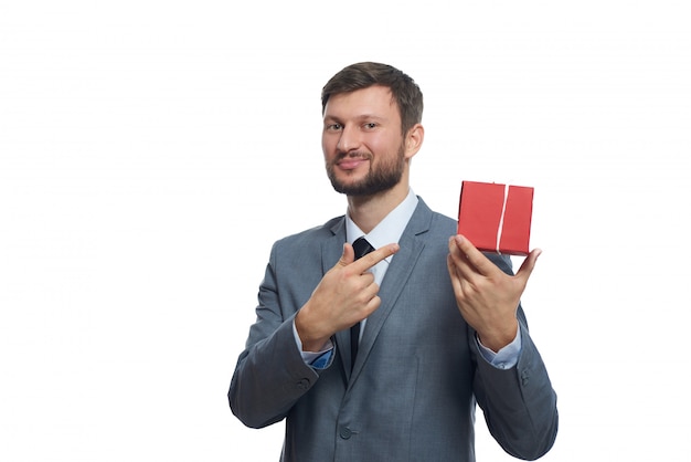 Porträt eines fröhlichen jungen Geschäftsmannes in einem Anzug, der ein kleines rotes Geschenk hält