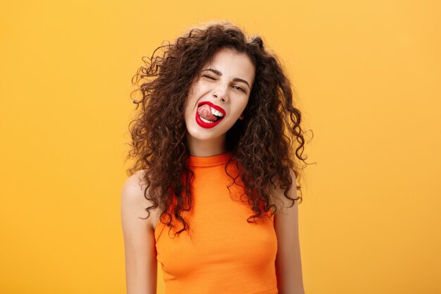 Porträt eines fröhlichen, verspielten und stilvollen kaukasischen Mädchens mit lockigem Haar und rotem Lippenstift, das zwinkert...