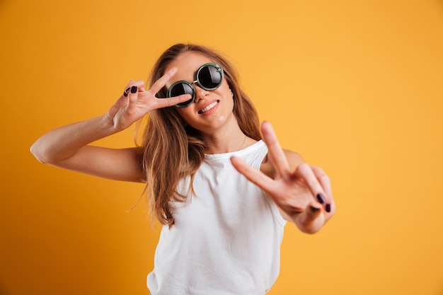 Porträt eines frohen jungen Mädchens in der Sonnenbrille, die Frieden zeigt