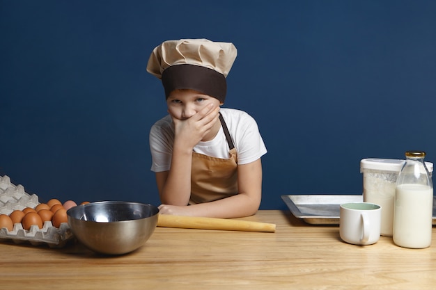 Porträt eines frustrierten 10-jährigen Jungen in Kochuniform, der den Mund bedeckt und sich verwirrt fühlt, während er zum ersten Mal selbst Pfannkuchen mit Milch macht