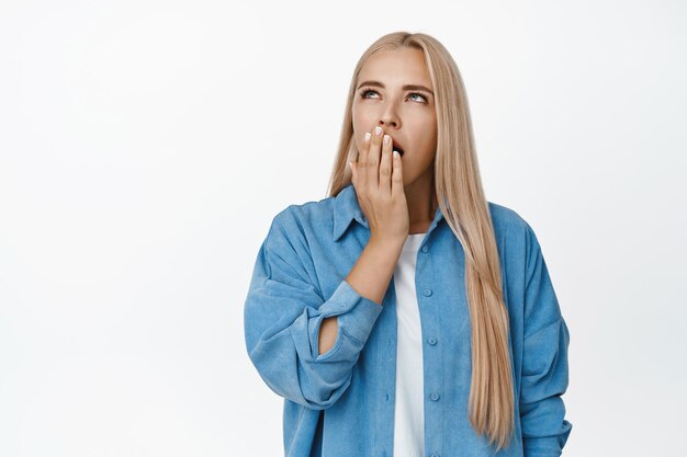 Porträt eines gähnenden blonden Mädchens, das den Mund mit der Hand bedeckt und gelangweilt nach oben schaut, das müde vor weißem Hintergrund steht