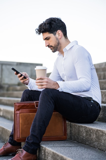Porträt eines Geschäftsmannes, der sein Handy benutzt, während er draußen auf Treppen sitzt