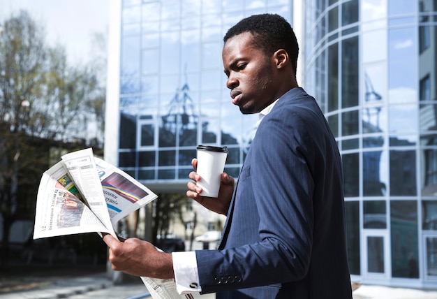 Porträt eines Geschäftsmannes, der vor dem Gebäude hält den Wegwerfkaffee liest die Zeitung steht