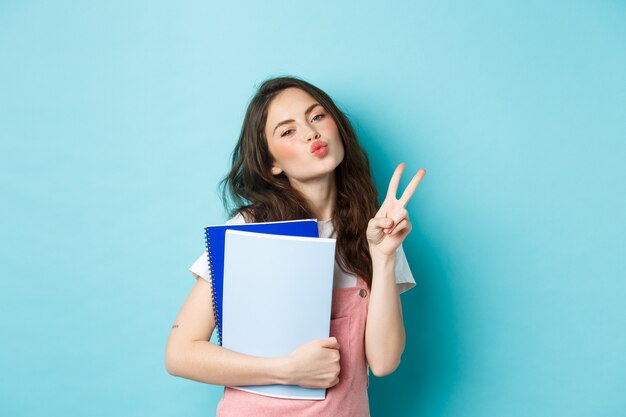 Porträt eines Glamour-Mädchens, das küssendes Gesicht und V-Zeichen zeigt, Notizbücher mit Hausaufgabenmaterial trägt und auf blauem Hintergrund steht