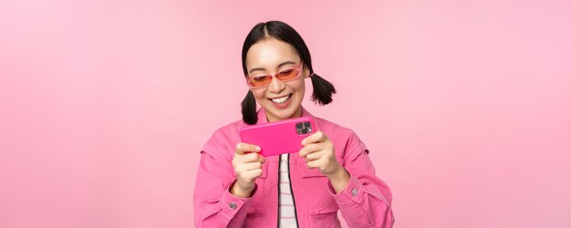 Porträt eines glücklichen asiatischen Mädchens, das auf dem Smartphone spielt und Videos in der Handy-App anschaut, die über rosa Hintergrund steht