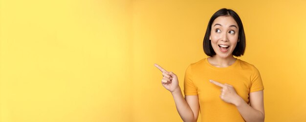 Porträt eines glücklichen asiatischen Mädchens, das mit dem Finger zeigt und nach links lächelt, erstaunt beim Auschecken des Promo-Banners, das Werbung vor gelbem Hintergrund zeigt