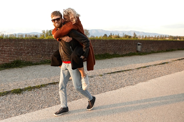 Porträt eines glücklichen attraktiven jungen bärtigen Kerls in den Schatten lächelnd breit froh, seiner schönen Freundin, die müde ist, huckepack fahren, kann nicht alleine gehen. Menschen, Liebe und Beziehungen