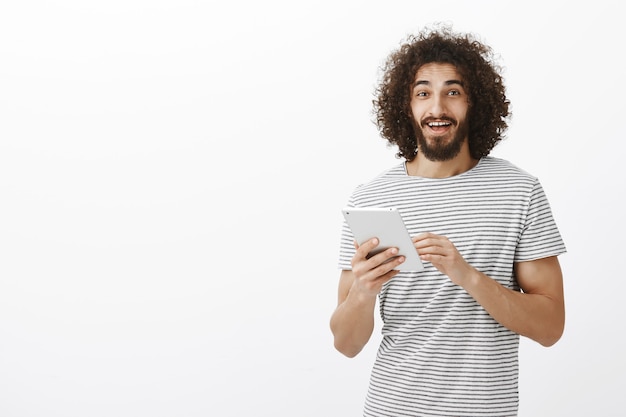 Porträt eines glücklichen freundlichen hispanischen bärtigen Mannes mit Afro-Frisur, der weiße digitale Tablette hält und breit auf Bildschirm anlächelt, positive Nachrichten mit Freunden teilt