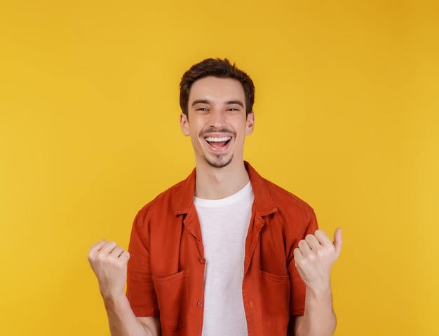 Porträt eines glücklichen, fröhlichen jungen Mannes, der dabei steht, Gewinnergeste zu ballen und die Fäuste zu ballen, die isoliert auf gelbem Wandhintergrundstudio bleiben