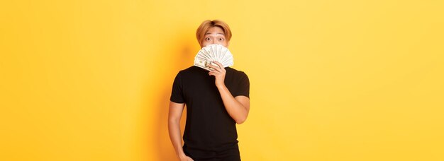 Porträt eines glücklichen, glücklichen Asiaten, der überrascht aussieht und Geld zeigt, das auf gelbem Hintergrund steht