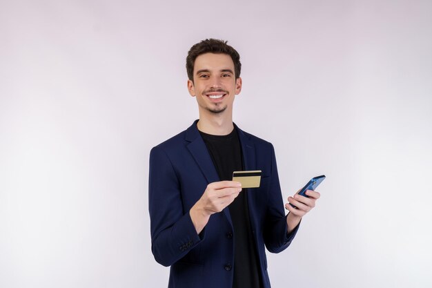 Porträt eines glücklichen jungen Geschäftsmannes, der mit einem Mobiltelefon steht und eine Kreditkarte hält, die auf einem Studio mit weißem Hintergrund isoliert ist