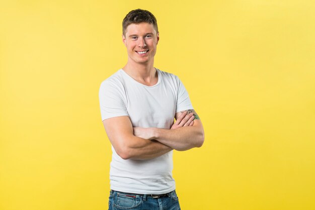 Porträt eines glücklichen jungen Mannes mit dem Arm kreuzte das Betrachten der Kamera, die gegen gelben Hintergrund steht