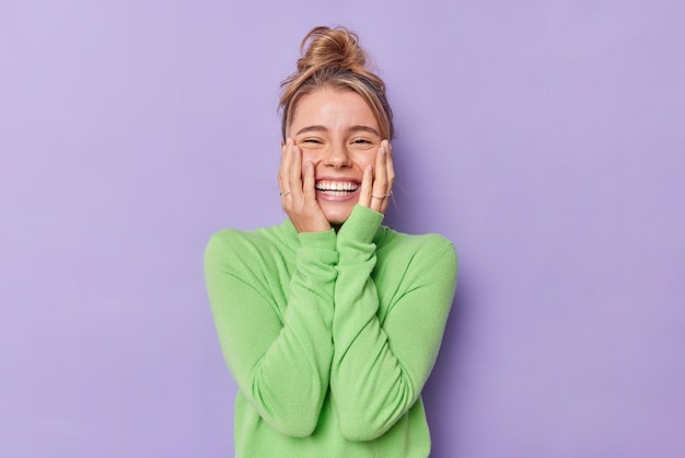 Kostenloses Foto porträt eines glücklichen jungen weiblichen modells hält die hände auf den wangen, lächelt breit, zeigt weiße zähne, fühlt sich froh, drückt aufrichtige gefühle aus, trägt einen grünen rollkragenpullover, der isoliert über violettem studiohintergrund steht
