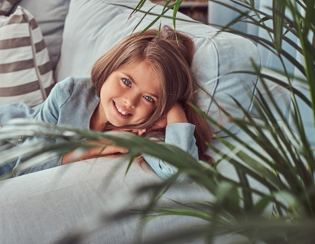 Porträt eines glücklichen kleinen Mädchens mit langen braunen Haaren und durchdringendem Blick, das auf eine Kamera blickt und allein zu Hause auf einem Sofa liegt