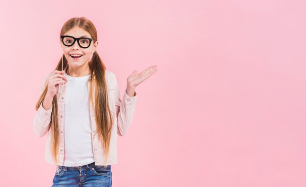 Porträt eines glücklichen Mädchens, das die Brillenstütze hält, zuckend gegen rosa Hintergrund