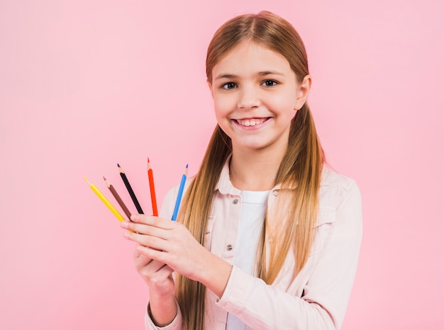 Porträt eines glücklichen Mädchens, das in der Hand farbige Bleistifte schaut, die zur Kamera gegen rosa Hintergrund schauen