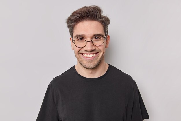 Porträt eines glücklichen Mannes mit dunklem Haar lächelt mit weißen Zähnen sieht selbstbewusst in die Kamera und trägt eine runde transparente Brille, schwarzes T-Shirt posiert im Studio vor weißem Hintergrund. Positive Gefühle