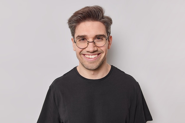 Porträt eines glücklichen Mannes mit dunklem Haar lächelt mit weißen Zähnen sieht selbstbewusst in die Kamera und trägt eine runde transparente Brille, schwarzes T-Shirt posiert im Studio vor weißem Hintergrund. Positive Gefühle