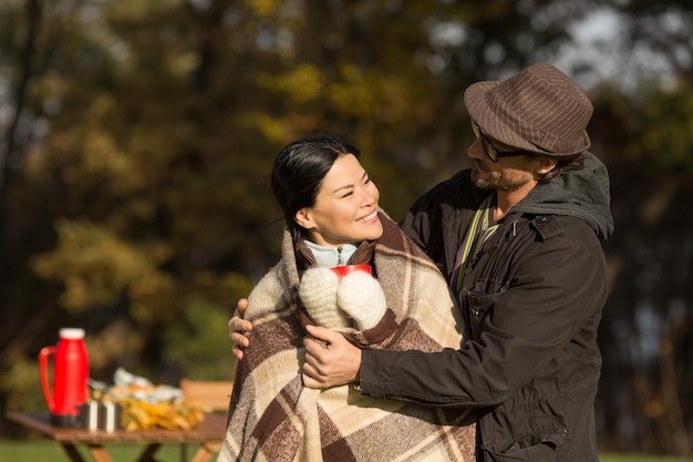 Porträt eines glücklichen Paares, das sich amüsiert Brünette Koreanerin, die ihren Ehemann anschaut, während er sie bei einem Picknick mit einem warmen Plaid bedeckt