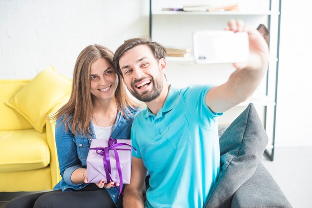 Porträt eines glücklichen Paars, welches das Valentinsgrußgeschenk nimmt selfie vom Smartphone hält