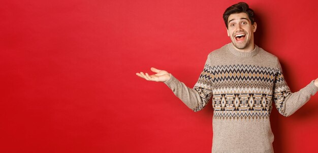Porträt eines glücklichen und überraschten gutaussehenden kaukasischen Kerls, der einen Weihnachtspullover trägt und die Hände seitlich ausbreitet und ahnungslos über rotem Hintergrund steht