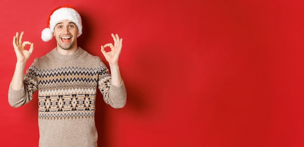 Porträt eines glücklichen und zufriedenen gutaussehenden Mannes, der einen Winterpullover und eine Weihnachtsmütze trägt, gute Zeichen zeigt, eine gute Weihnachtsfeier lobt und über rotem Hintergrund steht.