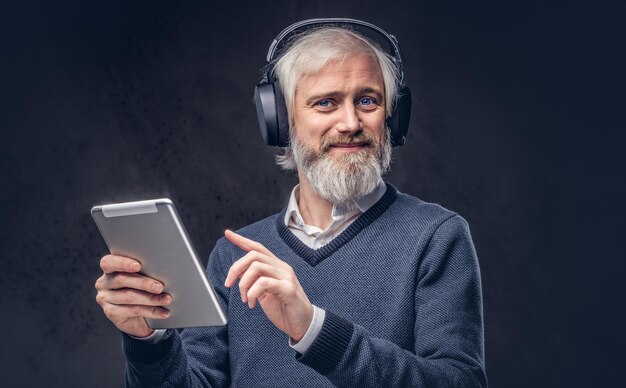 Porträt eines gutaussehenden älteren Mannes, der ein Tablet mit Kopfhörern in einem Studio vor dunklem Hintergrund verwendet.