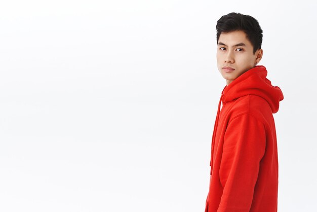 Porträt eines gutaussehenden asiatischen Hipster-Typen in rotem Hoodie, der im Profil steht und sich der Kamera mit entschlossenem, ernstem Ausdruck zuwendet, der auf weißem Hintergrund selbstbewusst steht