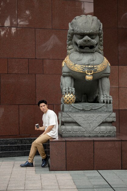 Porträt eines gutaussehenden asiatischen Mannes in der Stadt, der neben der Statue eine Tasse Kaffee trinkt