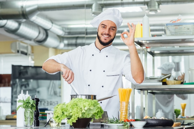 Porträt eines gutaussehenden bärtigen Chefkochs, der einen Topf hält und ein okayzeichen macht