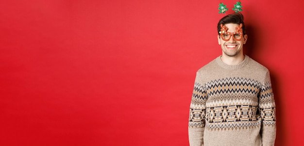 Porträt eines gutaussehenden bärtigen kerls mit partybrille und winterpullover, der erfreut lächelt, das neue jahr feiert, frohe weihnachten wünscht und über rotem hintergrund steht