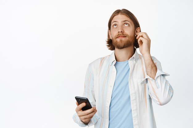 Porträt eines gutaussehenden blonden Mannes, der Kopfhörer in die Ohren steckt, mit zufriedenem Lächeln aufschaut, Smartphone in der Hand hält und auf Weiß steht