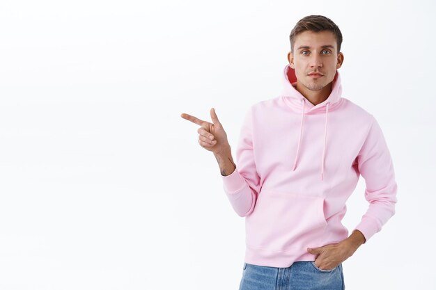 Porträt eines gutaussehenden blonden Mannes im rosa Hoodie, der mit dem Finger auf eine Leerstelle zeigt