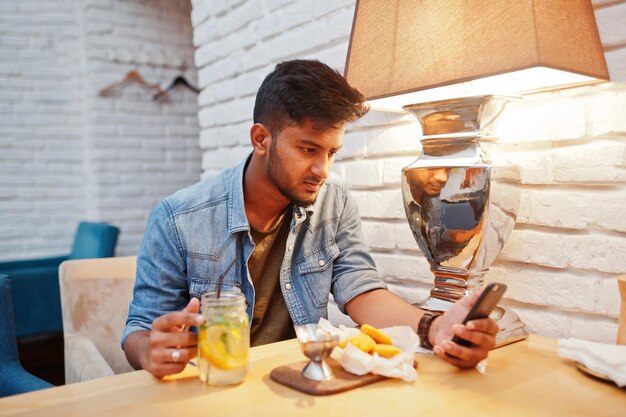 Porträt eines gutaussehenden erfolgreichen bärtigen südasiatischen jungen indischen Freiberuflers in blauem Jeanshemd, der im Café mit Chicken Nuggets und Limonade sitzt Halten Sie das Handy zur Hand