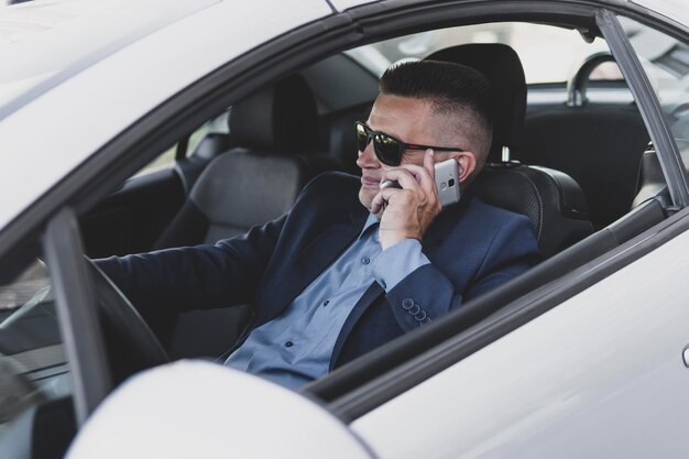 Porträt eines gutaussehenden geschäftsmannes, der telefonisch im sitz des autos sitzt und glücklich lächelt. fahrer kommuniziert während der fahrt auf dem smartphone. geschäftsmann, der fröhlich am steuer plaudert